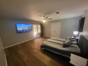 A bedroom with a bed, a television, a ceiling fan, and a door leading to a balcony.