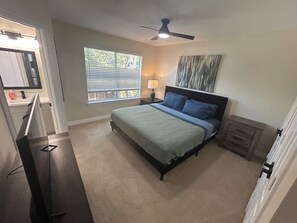 A bedroom with a bed, a ceiling fan, a window with blinds, a nightstand with a lamp, and a painting on the wall. - Enjoyable historic Duplex next door to Hutchins BBQ (McKinney)