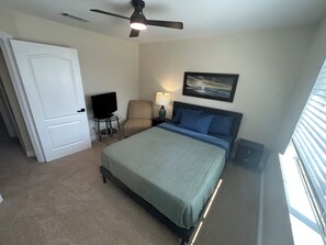 Quarto com cama, cadeira, televisão, ventoinha no teto e um quadro na parede.