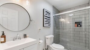 A modern bathroom with a round mirror, a white toilet, and a glass shower enclosure. - Charming 2BR Rear Unit Retreat Near Atlanta (Lithia Springs)