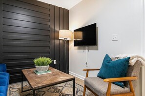 A modern living room with a dark wooden wall, a flat-screen TV mounted above a wooden coffee table with a potted plant, and a blue sofa. - Charming 2BR Rear Unit Retreat Near Atlanta (Lithia Springs)