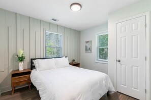 Ein Schlafzimmer mit einem Bett, zwei Nachttischen, einem Fenster mit Jalousien, einer Tür und einer kleinen Pflanze auf einem Nachttisch.