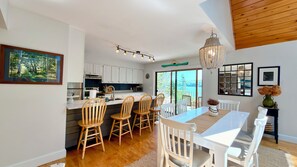 A modern kitchen with white cabinets, a dining area with wooden chairs, and a view of the outdoors. - Arden House | A Riverside Vacation Rental in Midcoast Maine (Cushing)