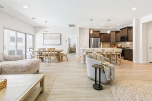 A modern living room with a sofa, a dining area with chairs, a kitchen with wooden cabinets, and a large window. - Mohican Escape | Stylish Backyard Retreat (Lake Havasu City)