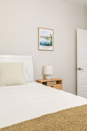 A hotel room with a bed, a nightstand with a lamp, a framed picture of Lake Havasu, and a white door. - Mohican Escape | Stylish Backyard Retreat (Lake Havasu City)