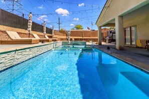 A swimming pool with lounge chairs and umbrellas, surrounded by a fence and buildings. - Mohican Escape | Stylish Backyard Retreat (Lake Havasu City)