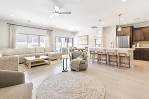Un salon moderne avec un canapé, une table basse et un tapis rond. On y trouve également un coin repas avec des chaises et une cuisine équipée d’un réfrigérateur et de placards.