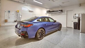 Ein blauer BMW M50 in einer sauberen, gut beleuchteten Garage mit einer weißen Tür, einer weißen Decke und einem an der Decke befestigten Projektor.