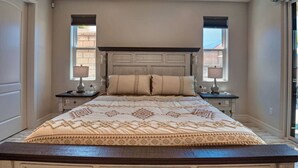 Ein Schlafzimmer mit einem großen Bett, zwei Nachttischen mit Lampen, einem dekorativen Kopfteil und Fenstern mit Jalousien.