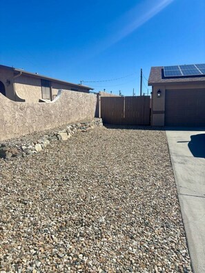 A residential area with a gravel driveway, a wooden fence, and a house with a solar panel on the roof. - Pet friendly home w/ pool, spa & toiletries (Lake Havasu City)