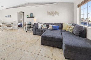 Un salon avec un canapé d’angle gris, une table à manger avec des chaises et une grande fenêtre donnant sur un beau paysage.