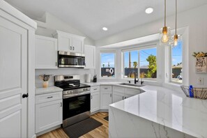 A modern kitchen with white cabinetry, stainless steel appliances, a large island, and a view of the outdoors. - Pool + Spa Retreat | Game Garage | Trailer Parking (Lake Havasu City)