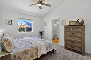 A bedroom with a bed, a dresser, a ceiling fan, a window with a view, and a door leading to a bathroom. - Pool + Spa Retreat | Game Garage | Trailer Parking (Lake Havasu City)