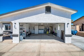 A single-story building with a garage entrance, a visible kitchen area, and a patio with outdoor seating. - Pool + Spa Retreat | Game Garage | Trailer Parking (Lake Havasu City)