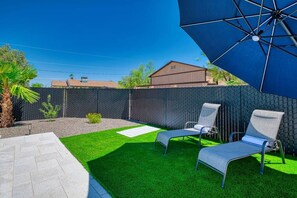 A backyard with artificial grass, two lounge chairs, a patio table, and a large blue umbrella. - Pool + Spa Retreat | Game Garage | Trailer Parking (Lake Havasu City)