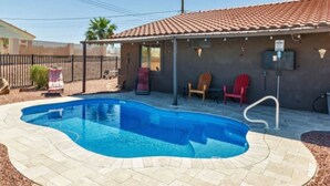 Une piscine avec une terrasse carrelée, entourée d’une clôture, et un bâtiment avec un toit en tuiles en arrière-plan.