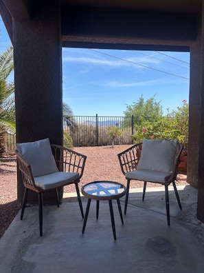 Two wicker chairs with cushions, a small round table, and a view of a garden with a fence and plants. - Pet-Friendly | Sunset View Deck | Pool & Spa (Lake Havasu City)