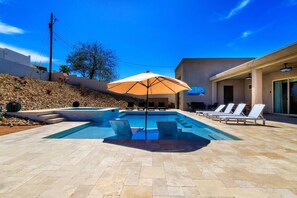 A swimming pool with lounge chairs and an umbrella, surrounded by a landscaped area with steps and a building in the background. - Resort-Style Entertainer | Near Launches (Lake Havasu City)