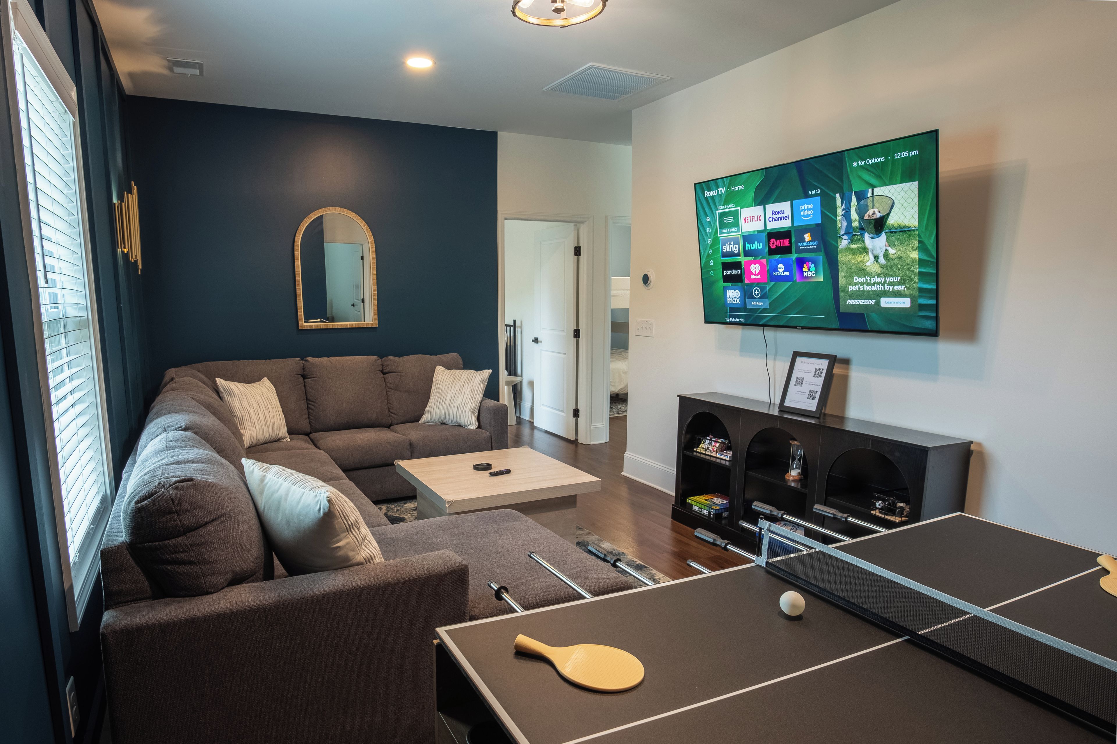 Una sala de estar con un sofá seccional, una mesa de ping pong y un televisor de pantalla plana montado en la pared.