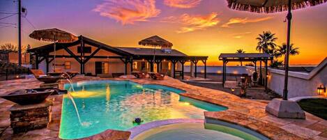 A swimming pool with lounge chairs and umbrellas, surrounded by buildings with a sunset view.
