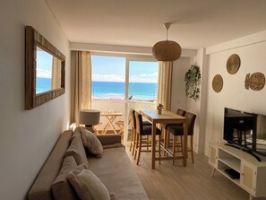A living room with a sofa, dining table, chairs, a TV, and a view of the ocean. - Sea View Apartment - Right on the Beach in Costa da Caparica (Costa de Caparica)