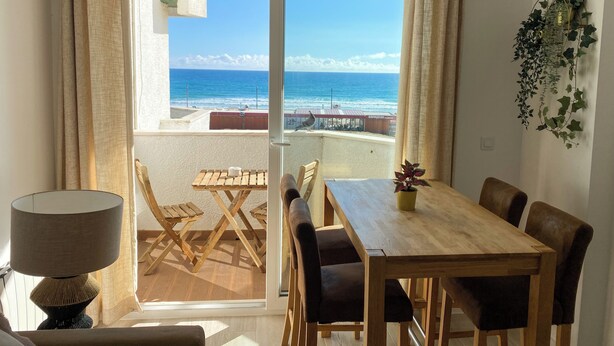 Un balcón con una mesa y sillas de madera, vistas al océano y una planta en maceta en la pared.