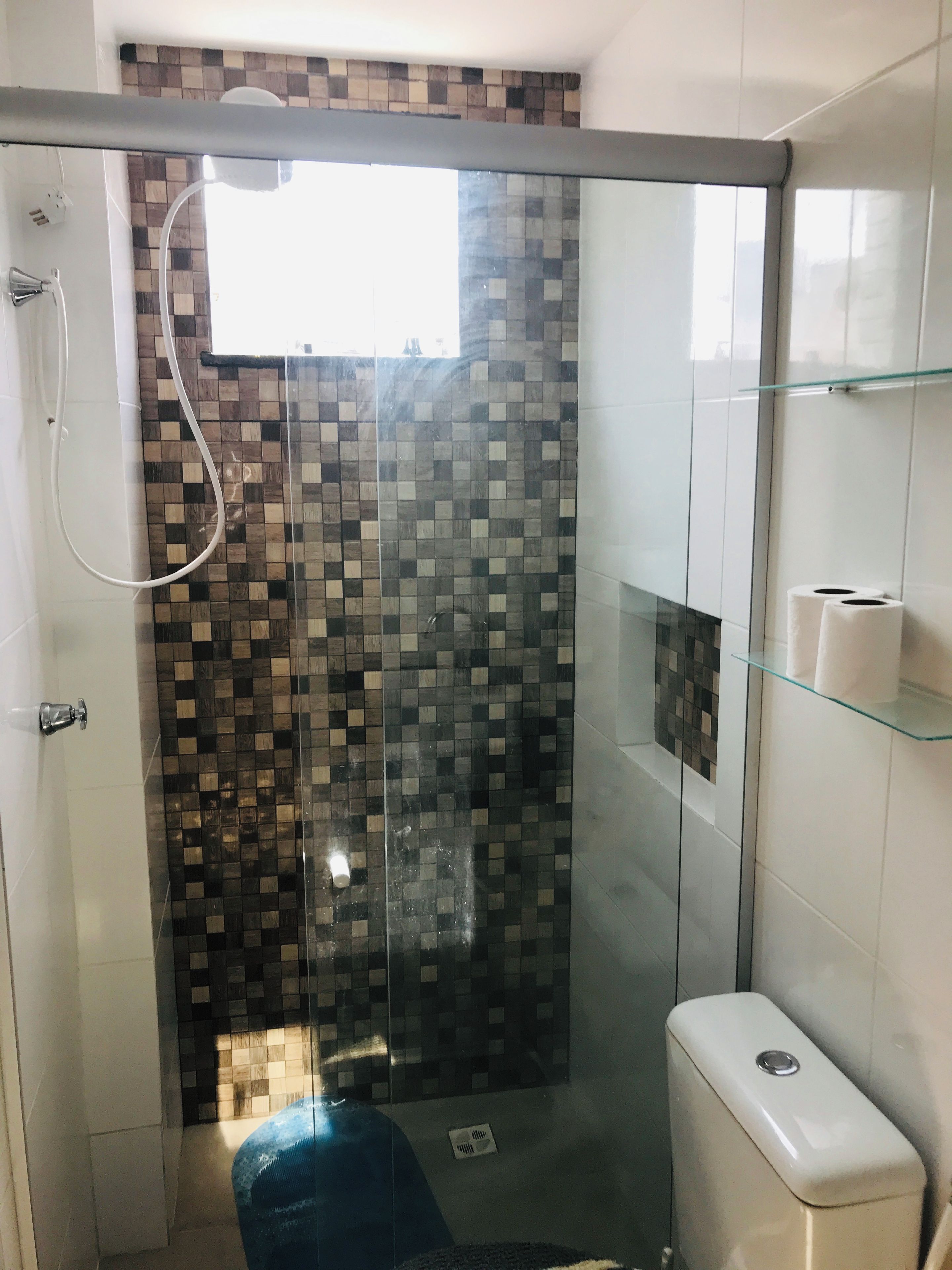 A shower with a glass door, a tiled wall, and a glass shelf with a toilet paper holder.