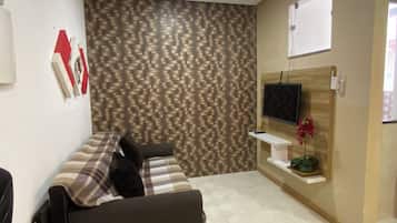 Ein Zimmer mit einem Wandmuster, einem an einer Holzplatte montierten Flachbildfernseher, einem weißen Regal mit einer Topfpflanze und einem sofaartigen Sitzmöbel mit Karomuster.