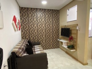 A room with a patterned wall, a flat-screen TV mounted on a wooden panel, a white shelf with a potted plant, and a plaid-patterned sofa. - Apto 101 (Belém)