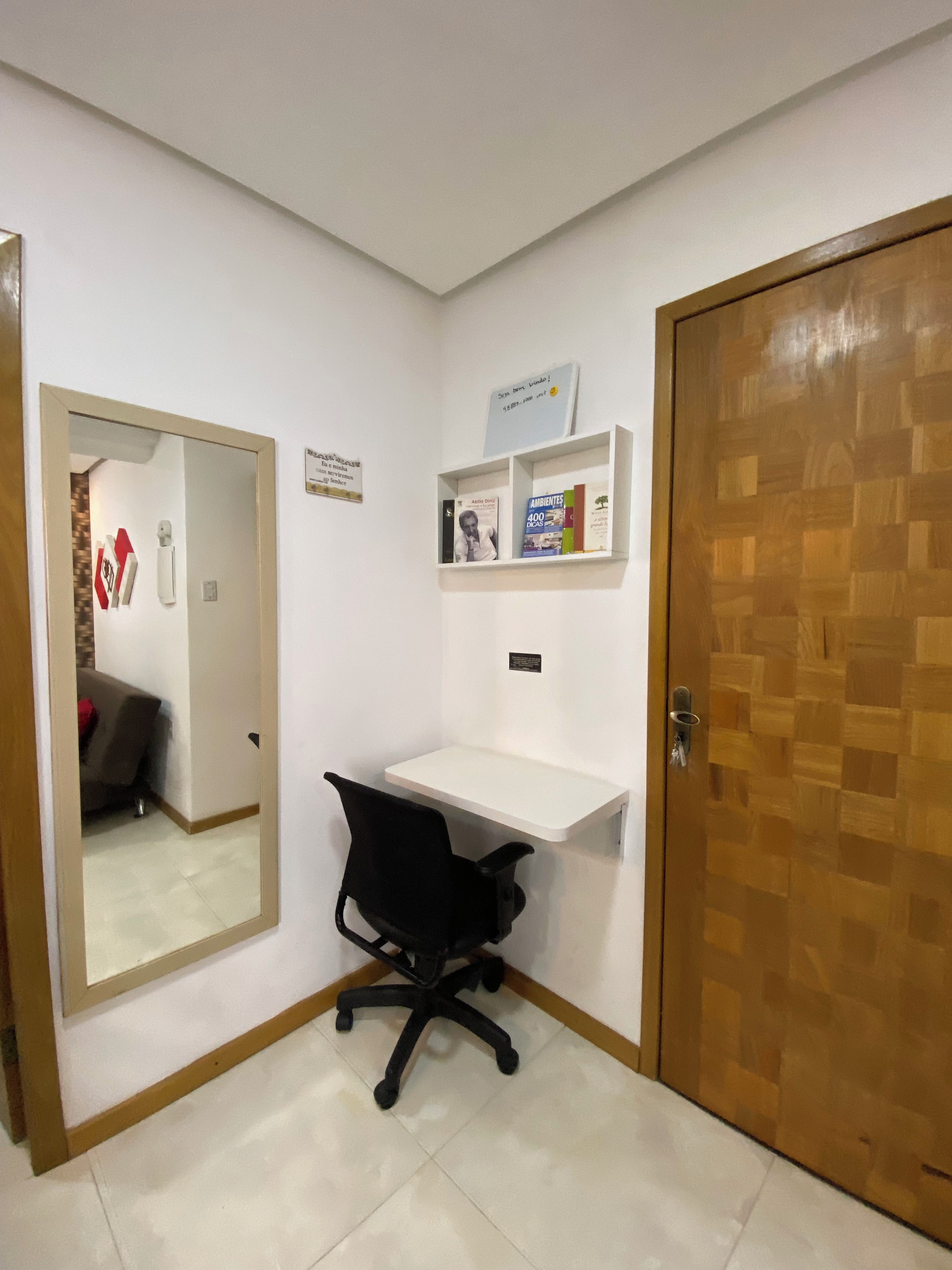 A small office space with a desk, chair, and a mirror. There is a wooden door and a shelf with books and a sign.