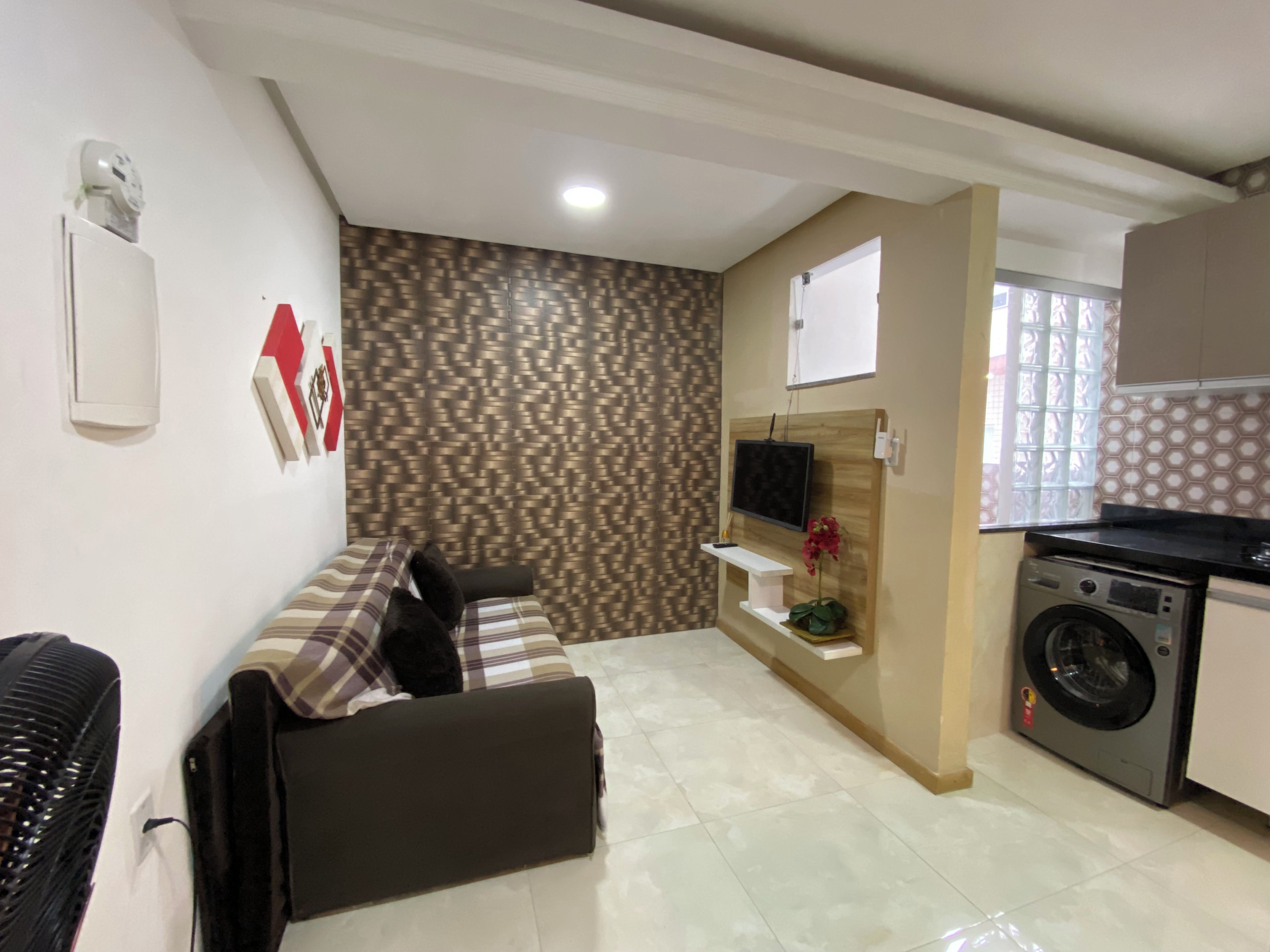 A living room with a sofa, a television mounted on the wall, and a washing machine in the adjacent kitchen area.