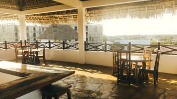 An outdoor dining area with tables and chairs, a thatched roof, and a view of a cityscape.