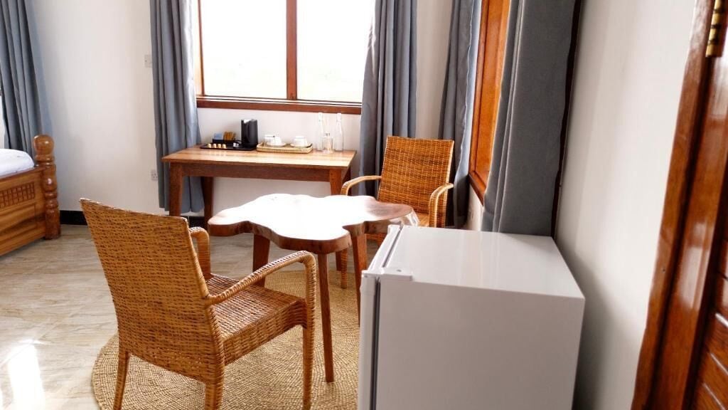 Una habitación con cama, una mesa de comedor, dos sillas de mimbre, un refrigerador y una ventana con cortinas.