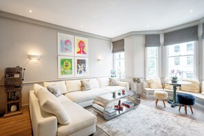 Ein modernes Wohnzimmer mit einer Eckcouch, einem Couchtisch und einem runden Tisch mit Hockern. An der Wand befinden sich gerahmte Kunstwerke und ein Kamin mit Sims.