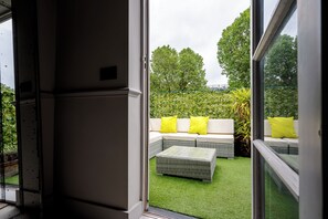 Vue depuis l’intérieur d’une pièce sur un espace extérieur aménagé avec une pelouse artificielle verte, un canapé en osier avec des coussins jaunes et une plante en pot.