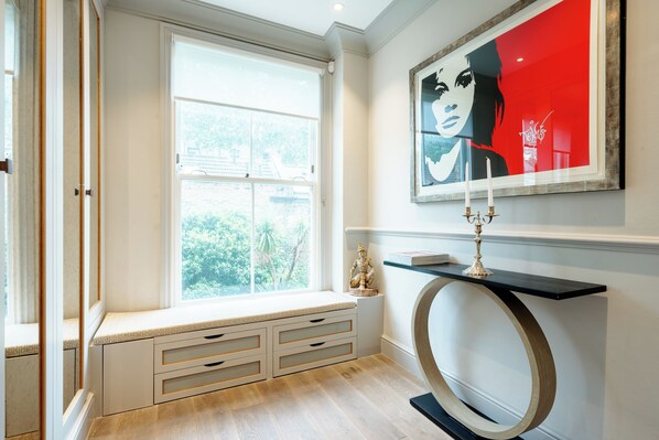 Une pièce avec un banc intégré près d’une grande fenêtre, une œuvre d’art encadrée au-dessus d’une console, et une statue décorative posée sur la console.