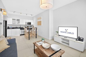 Een moderne woonkamer met een televisie waarop 'MONTE CARLO By MiamiVacations' wordt weergegeven, een eethoek met een houten tafel en stoelen, en een keuken met witte kasten en apparatuur.