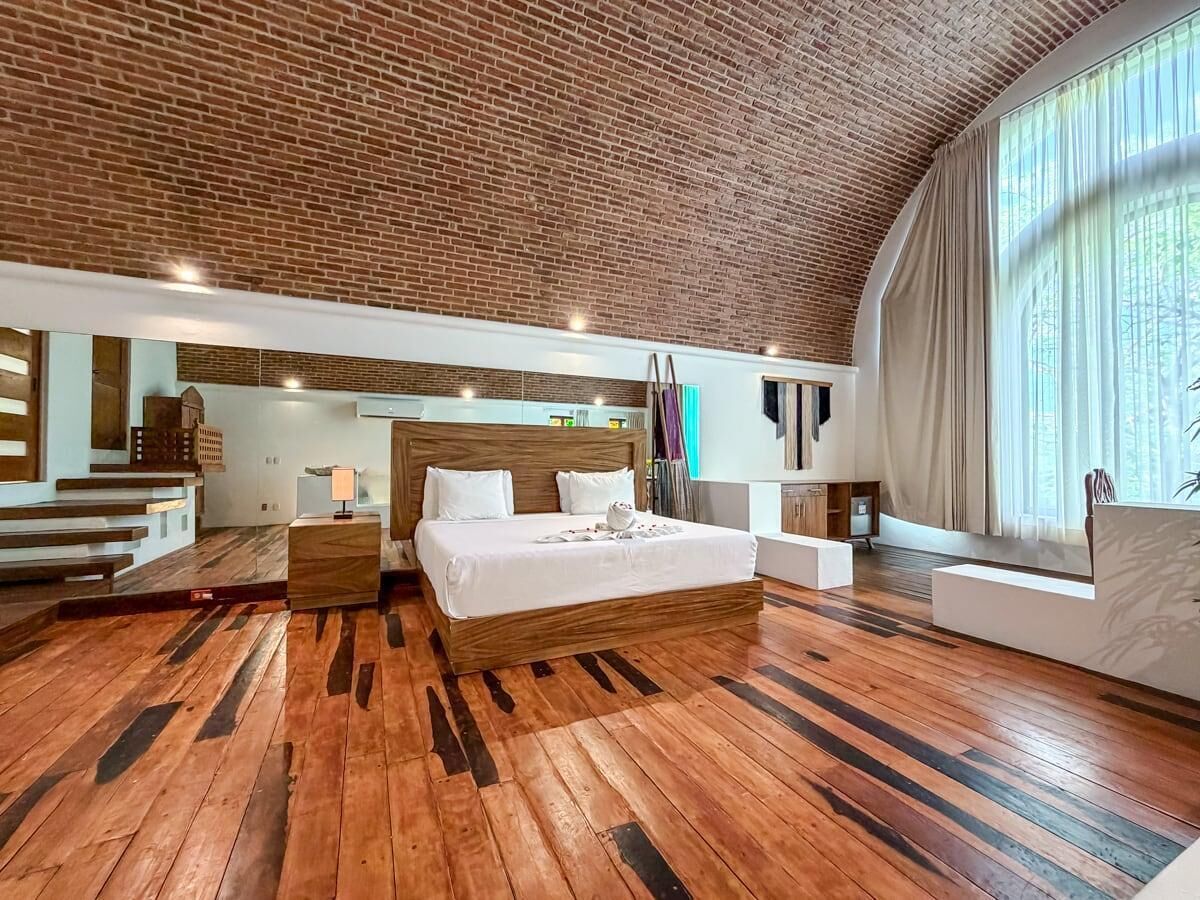 A spacious bedroom with a large bed, wooden flooring, a brick ceiling, and a large window.