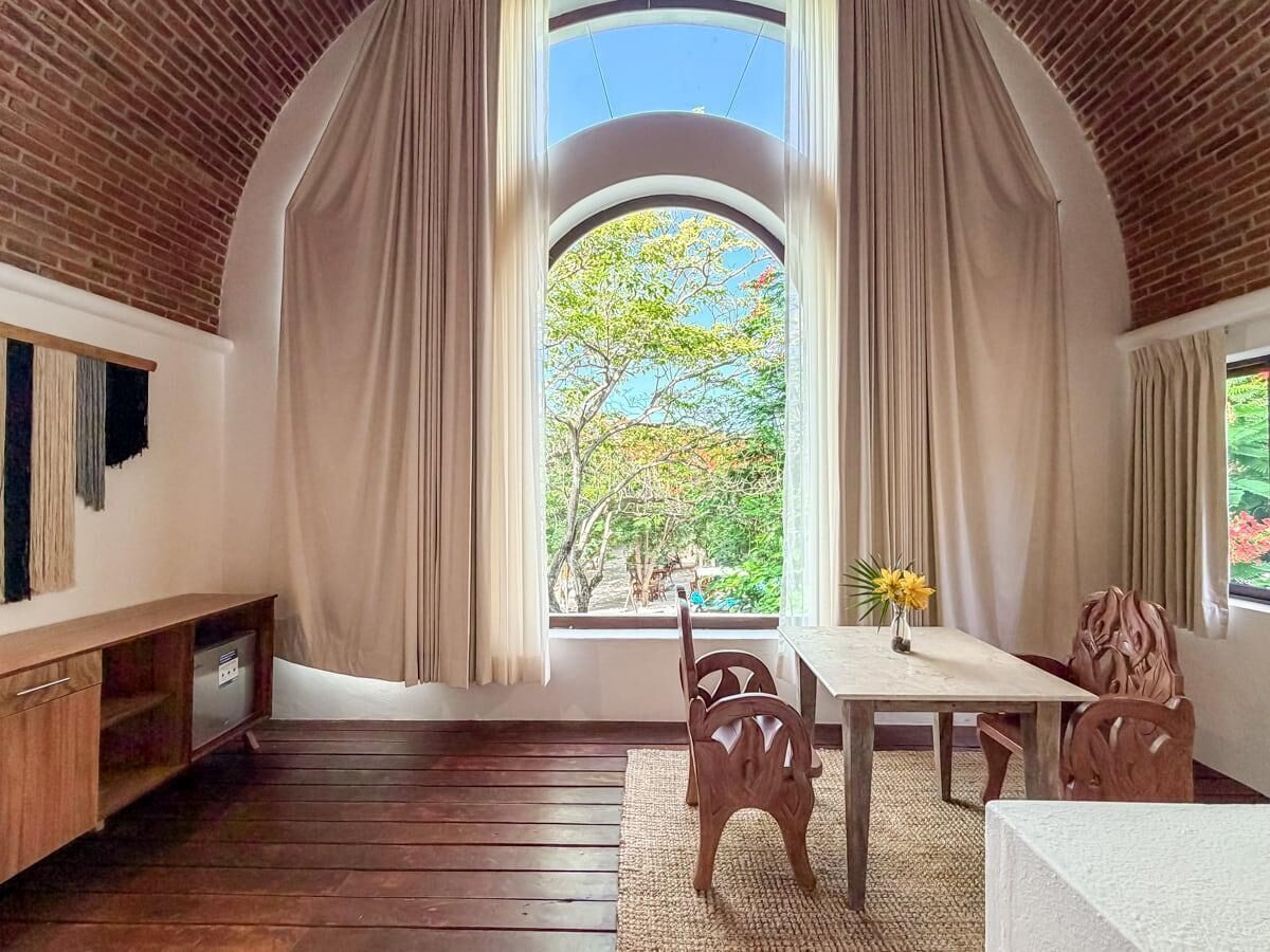 A room with a wooden floor, a dining table with chairs, a TV on the wall, and a window with curtains.