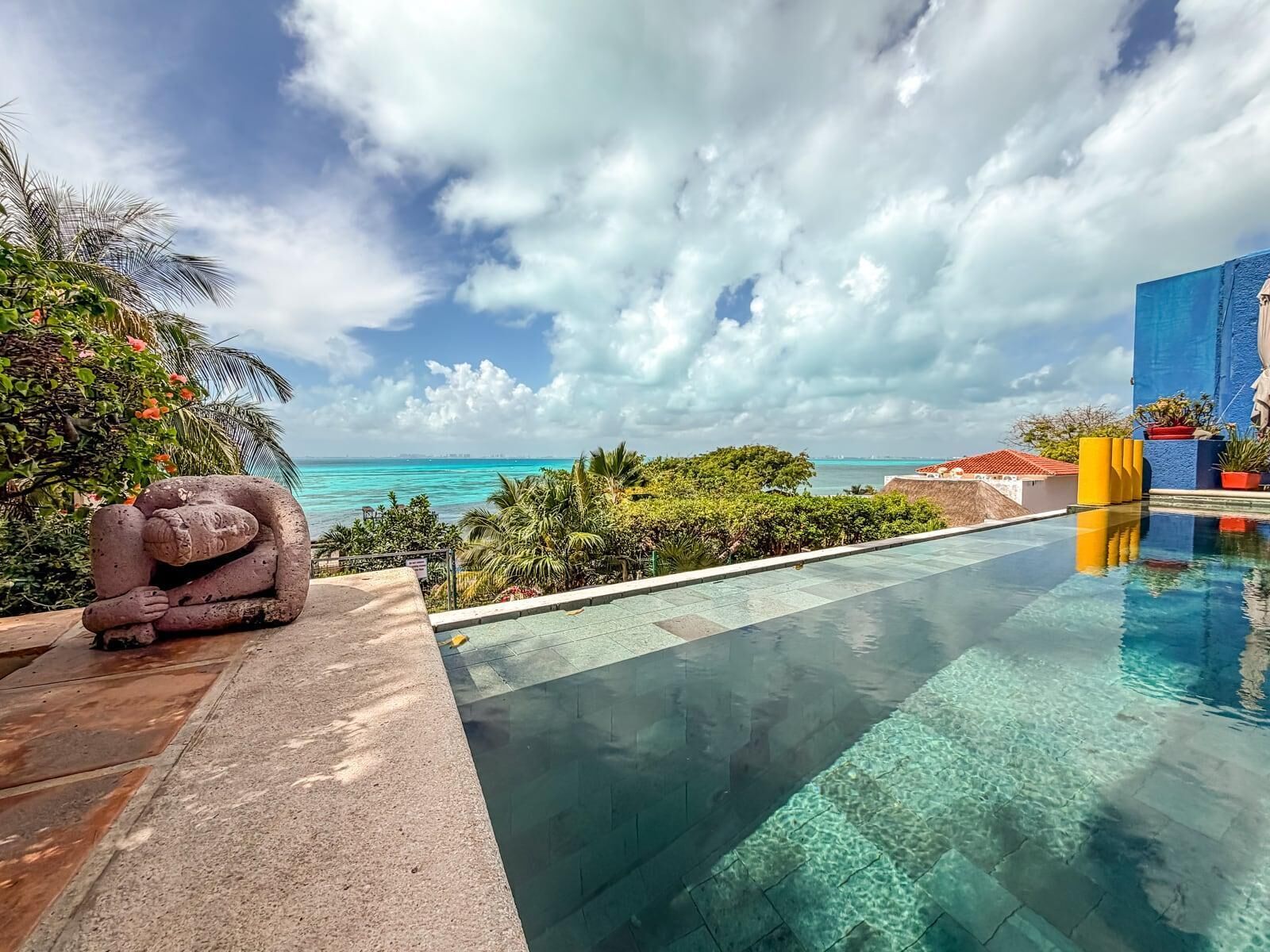 A clear pool with a view of the ocean, a stone sculpture, and tropical vegetation.