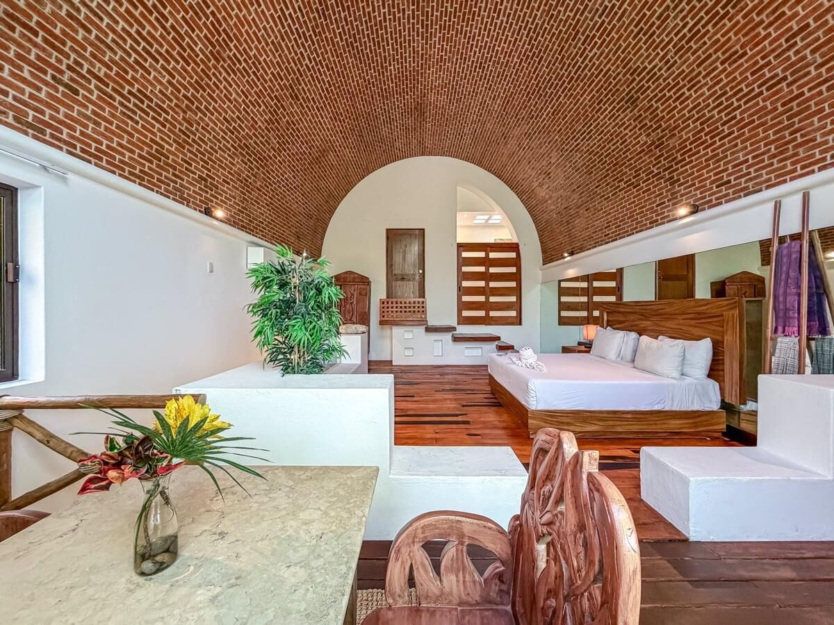 A modern interior with a brick ceiling, a central seating area, a bed, and a dining table with a vase of flowers.