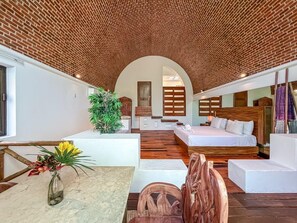 A modern interior with a brick ceiling, a central seating area, a bed, and a dining table with a vase of flowers. - Junior Suite Garden View at Casa de los Suenos (Isla Mujeres)