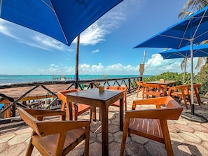 A beachside restaurant with wooden tables and chairs, blue umbrellas, and a view of the ocean. - Junior Suite Garden View at Casa de los Suenos (Isla Mujeres)