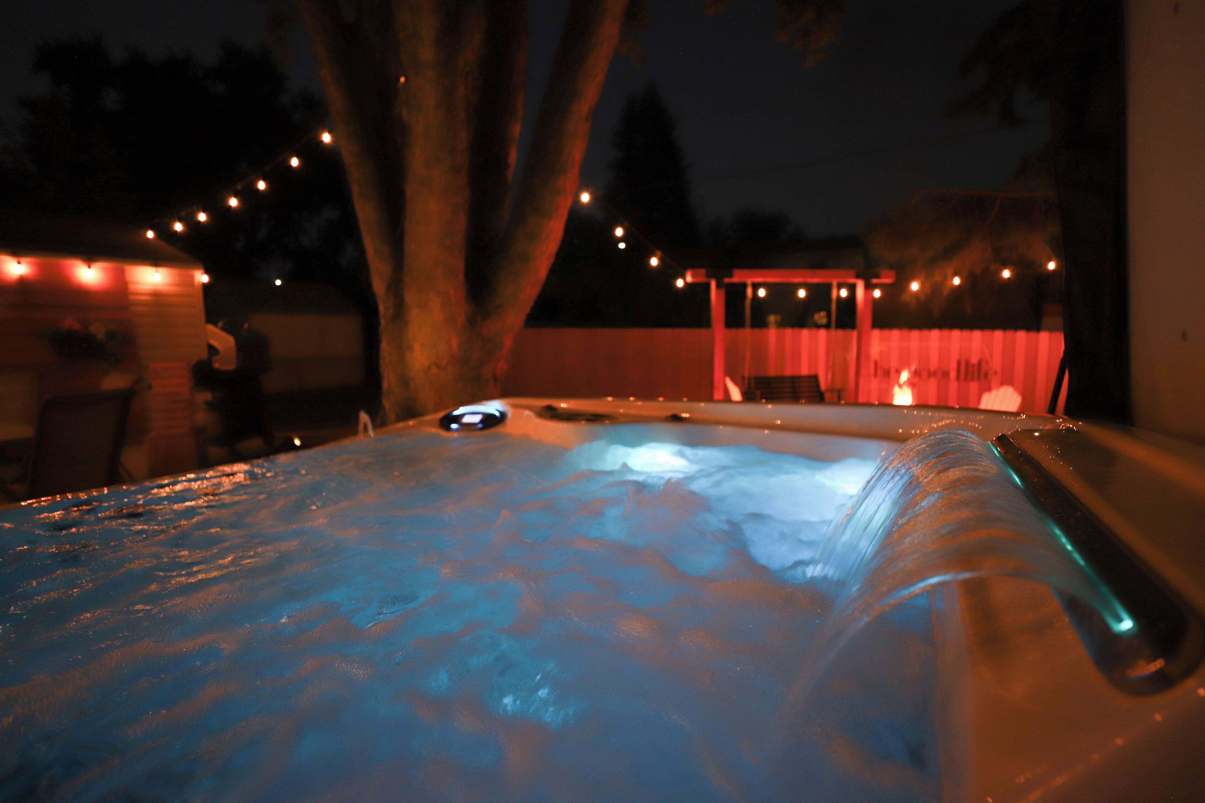 A hot tub with a waterfall feature, surrounded by string lights and a covered seating area.