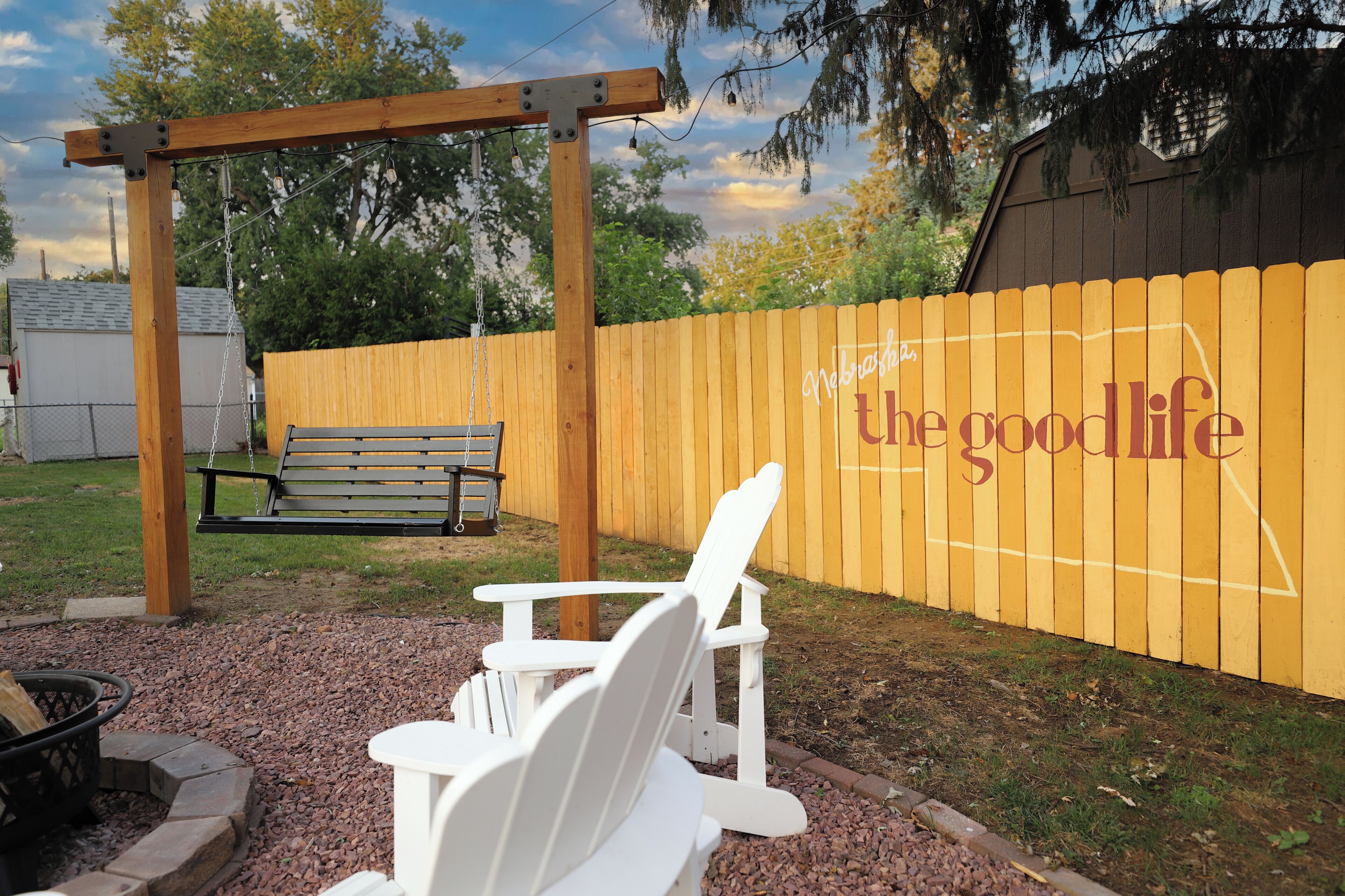 Een houten pergola met een schommel, een bank en Adirondack-stoelen in een achtertuin met een houten hek en een muur waarop 'Nebraska the good life' staat geschilderd.
