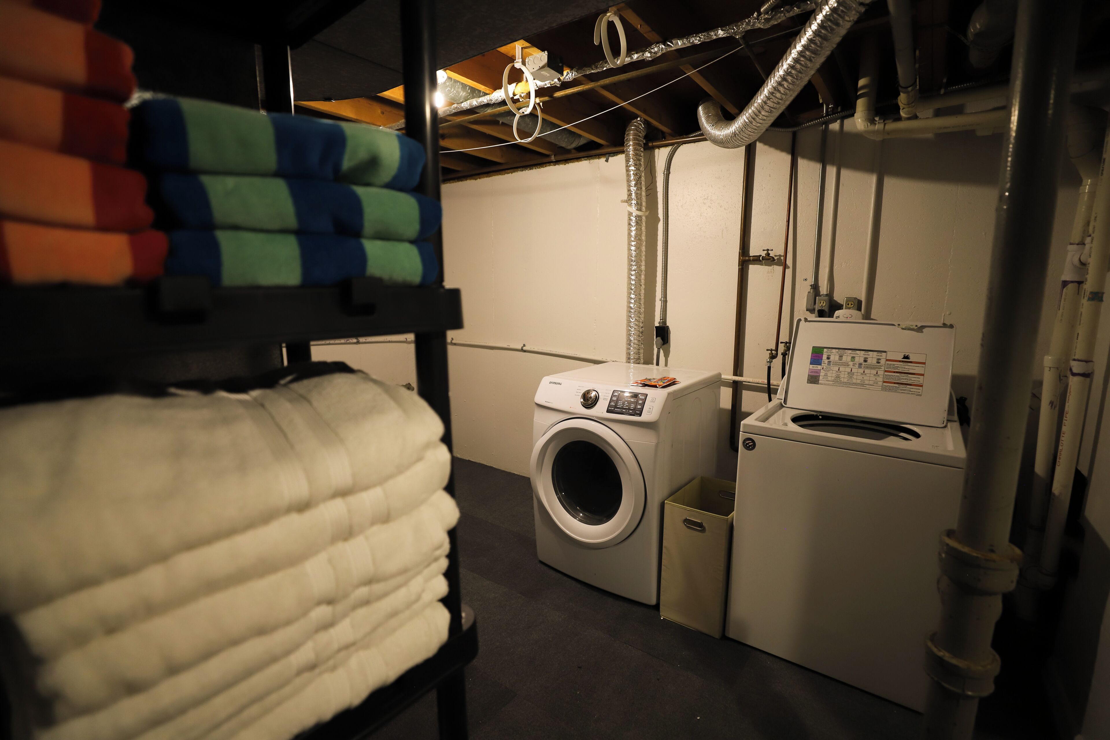 Een wasruimte met een wasmachine en droger, een stapelbed met kleurrijk beddengoed en zichtbare leidingen.