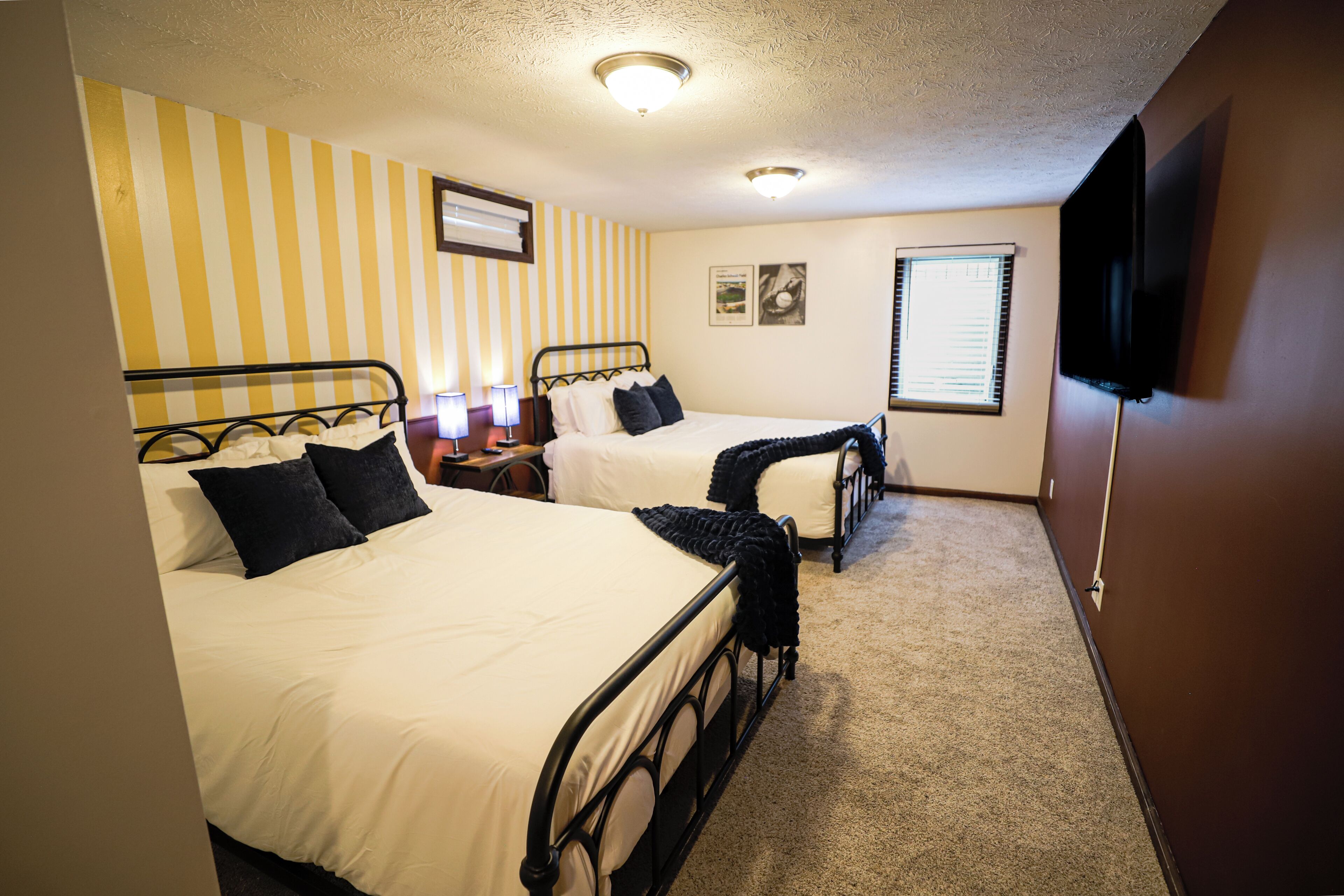 A hotel room with two single beds, a flat-screen TV mounted on the wall, and a window with blinds.