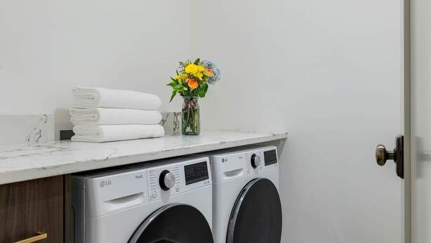 A laundry room with two front-loading washing machines, a marble countertop, and a vase with flowers. - The Bloomberg at Bloomberg Suites (Chanhassen)