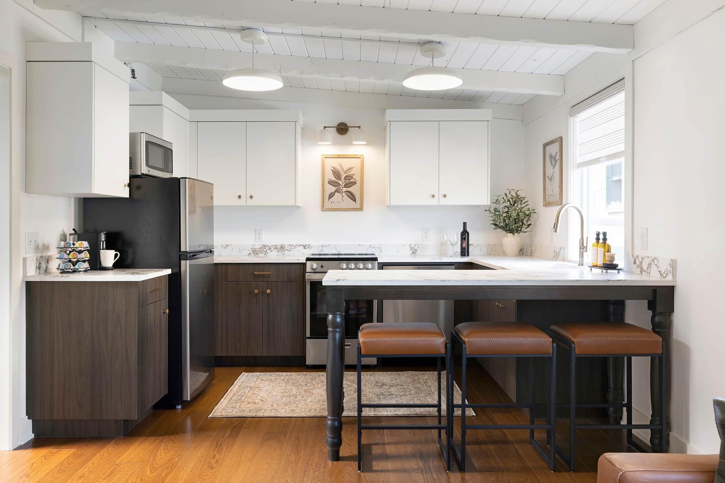 Eine moderne Küche mit dunklen Holzschränken, einem Edelstahlkühlschrank, einem eingebauten Backofen und einer Marmorarbeitsplatte. Drei Barhocker und ein großes Fenster sorgen für natürliches Licht.