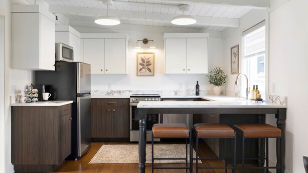Cocina moderna con armarios de madera oscura, refrigerador de acero inoxidable, horno integrado y encimera de mármol. Hay tres taburetes de bar y un ventanal que proporciona luz natural.
